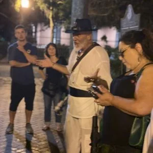 Tour guide in period costume with wide-brimmed hat gestures while speaking to group of tourists on cobblestone street at night