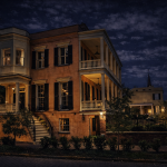 A two story historic home stands beneath a star filled night sky, its warm interior lights glowing through tall windows framed by dark shutters. The structure features a wraparound balcony with white balustrades, a columned porch, and a graceful curved staircase leading to the entrance. Soft lamplight casts gentle shadows across the stucco facade, while nearby trees and an iron fence add depth and texture to the scene. A church steeple rises faintly in the background, enhancing the quiet, atmospheric mood of this elegant Southern residence.