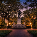 Johnson Square in historic Savannah, Georgia, at dusk with monument and glowing gas lamps beneath moss draped oak trees, a legendary stop on Savannah ghost tours and haunted Savannah walking tours.