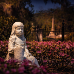 Twilight photograph of a white marble statue of a young girl seated among vibrant pink azaleas in a historic Savannah cemetery, with an iron fence and softly lit headstones visible in the background beneath moss-draped trees.