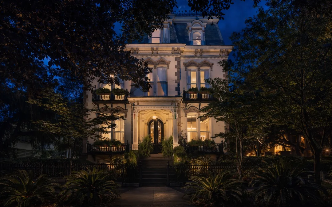 Victorian mansion illuminated at night with ornate white facade, wrought iron balconies, and lush garden framed by trees