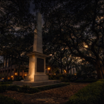 Nighttime view of a tall stone obelisk monument in a Savannah square, surrounded by oak trees and brick walkways. Warm lamplight glows through the trees, casting soft shadows across the landscaped grounds and nearby buildings.
