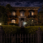 Nighttime view of a grand historic brick mansion in Savannah, featuring tall arched windows, wrought iron balconies, and a columned entrance. The home is warmly lit from within, framed by manicured hedges and an iron fence beneath a cloudy evening sky.