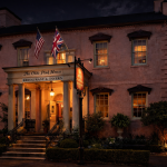 Nighttime view of The Olde Pink House Restaurant and Tavern in Savannah, a historic pink Georgian mansion with white columns, warmly lit windows, and American and British flags above the entrance. The front steps and landscaping are softly illuminated against a dark evening sky.