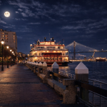 A moonlit view along Savannah’s River Street. Brick pavement stretches beside the dark water of the Savannah River, lined with glowing lampposts. A brightly lit riverboat is docked along the waterfront, its warm lights reflecting on the water. In the distance, the Talmadge Memorial Bridge spans the river beneath a full moon and scattered clouds, with port cranes and ships visible on the horizon.