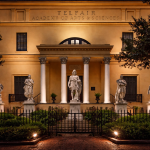 Evening view of the Telfair Academy of Arts and Sciences in Savannah, a historic neoclassical building with tall white columns, statues lining the front steps, and warm exterior lighting illuminating the façade behind an iron gate and brick walkway.
